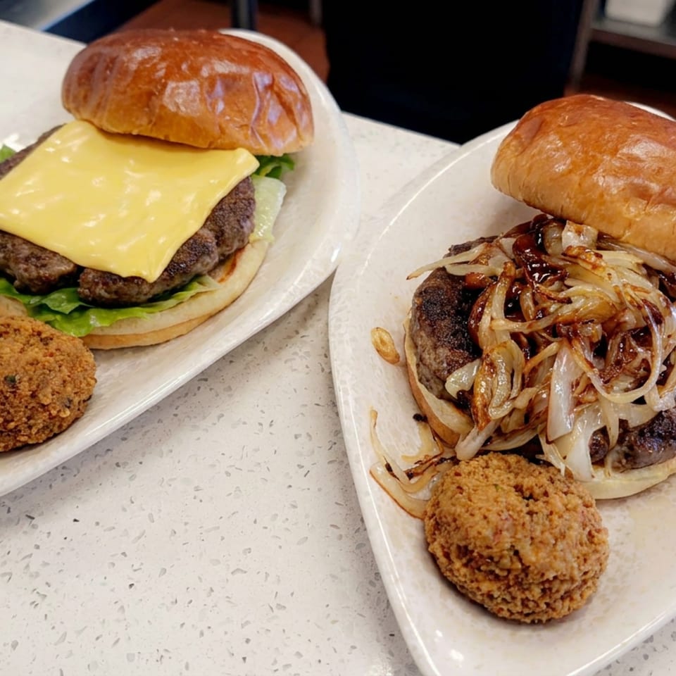 Two cheeseburgers on plates, one topped with lettuce and cheese and the other with grilled onions, each served with a fried croquette.