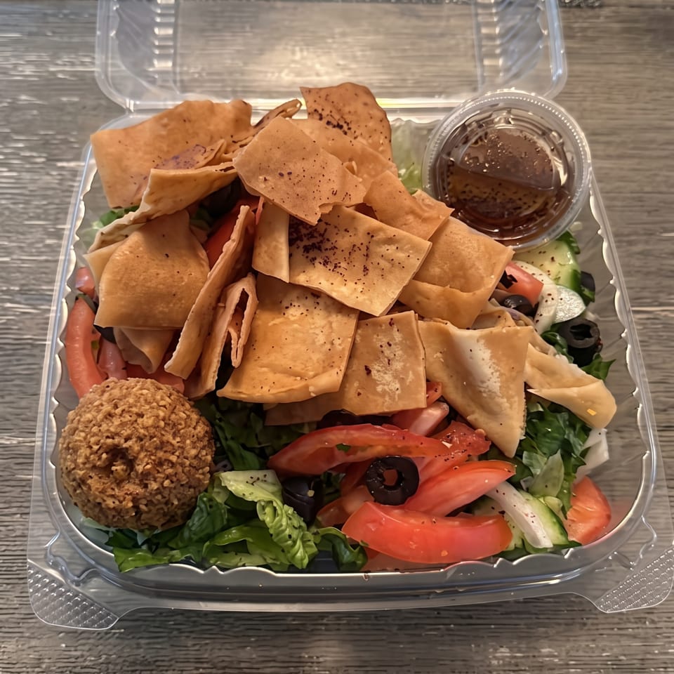 Open takeout container filled with chopped salad, pita chips, falafel, vegetables, and a cup of dressing.